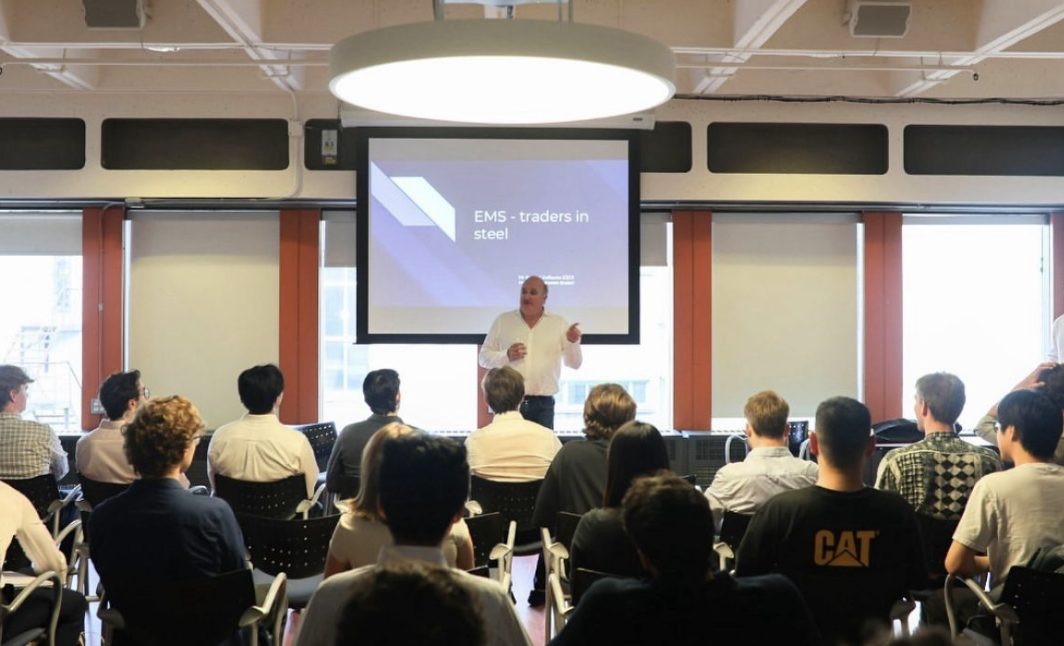 An MCTG speaker session on steel trading, with a presenter addressing seated members in front of a projected title slide